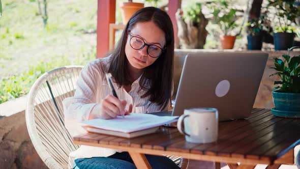 A Young Woman Freelancer Makes Notes in a Notebook and a Laptop