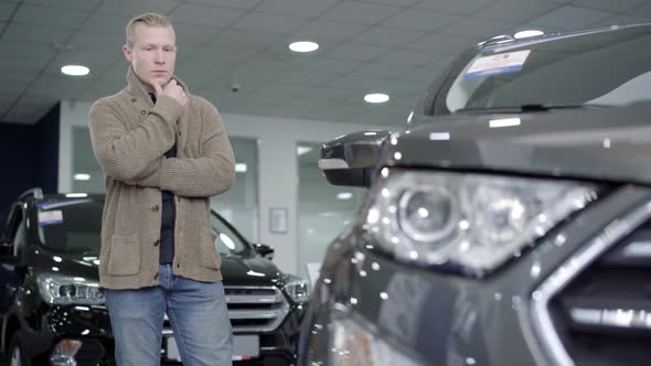 Portrait of Thoughtful Young Caucasian Man Choosing Automobile in Dealership or Showroom. Serious