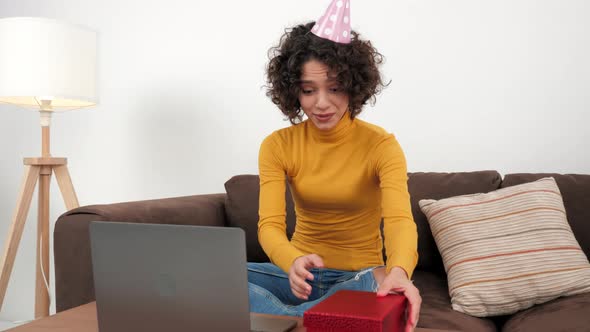 Happy Woman in Party Hat Opens Gift Box and Surprised Sitting in Couch at Home