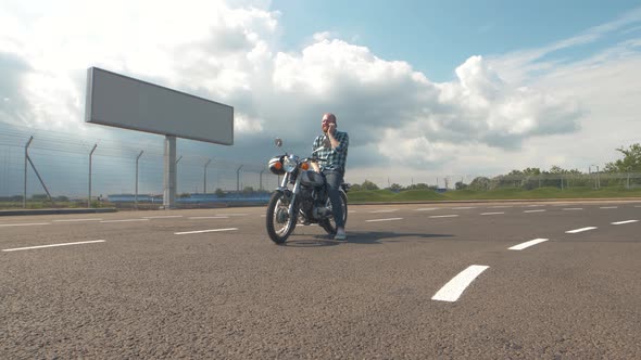 Young Man Motorcyclist with Smart Phone in Hands on Custom Motorcycle on Street