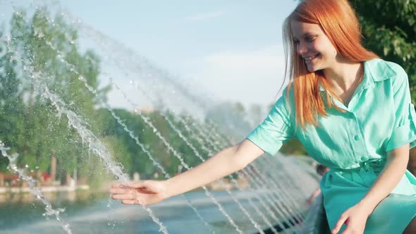 girl with red hair touches a hand stream of water from fountain, slow motion