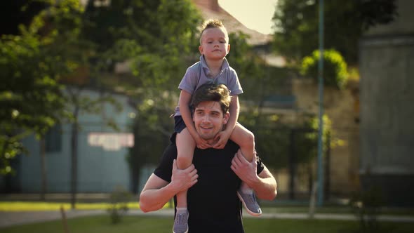 Young Dad in Sportswear is Smiling and Carrying Tired Kid Boy on His Shoulders Showing Something to