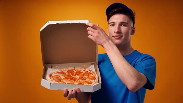 Young Deliveryman in Blue Uniform Opens a Box of Fresh Pizza Against Yellow Background