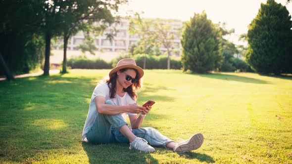 A Young Woman Uses Her Phone While Sitting on a Green Bright Lawn