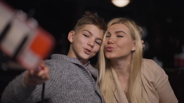 Smiling Caucasian Mother Gesturing Peace Taking Selfie with Teenage Son in Restaurant