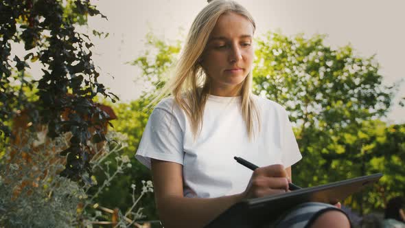 Young Girl is Sitting on a Bench of City Park and Drawing on a Digital Tablet with Stylus