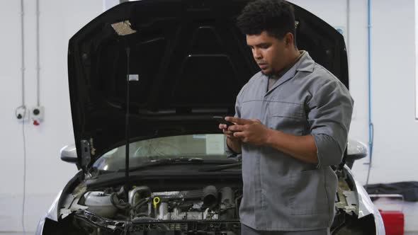 African American male car mechanic looking at an open car engine and talking on a smartphone