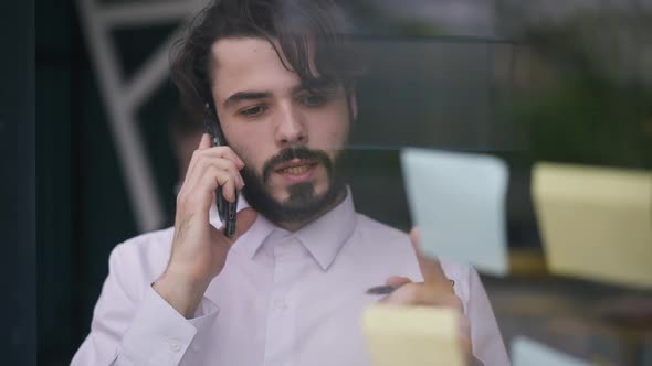 Portrait of Focused Young Bearded Caucasian Man Talking on Phone Writing on Adhesive Notes Standing