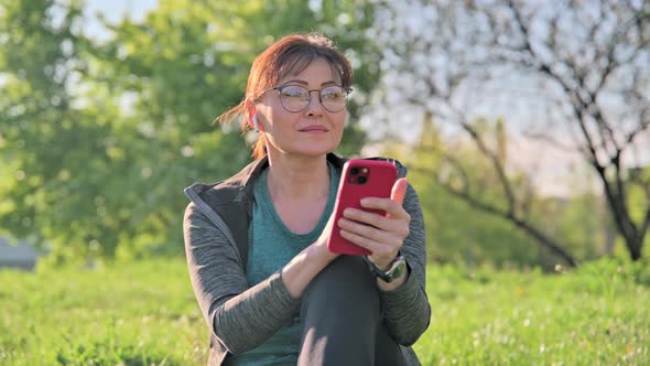 Mature Female in Headphones with Smartphone Listening Music Sitting on Grass