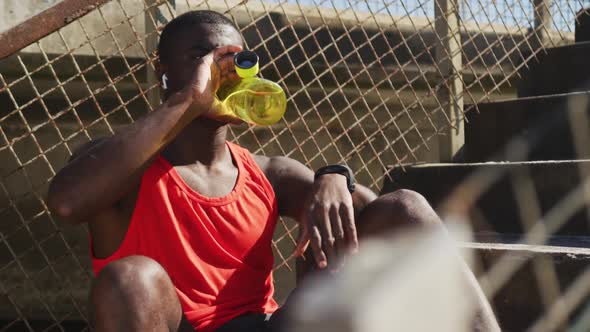 Tired african american man sitting, drinking from water bottle, taking break in exercise outdoors