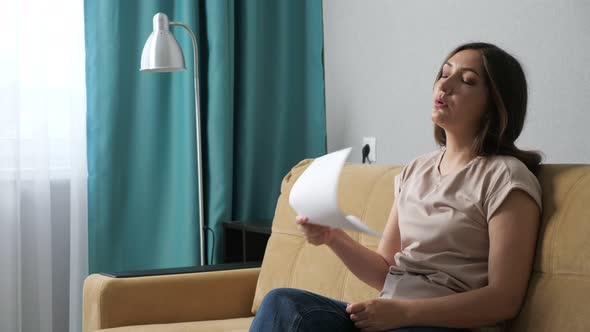 Side View of Young Woman Suffering From Fever Fanning with Sheet of Paper While Sitting on Sofa