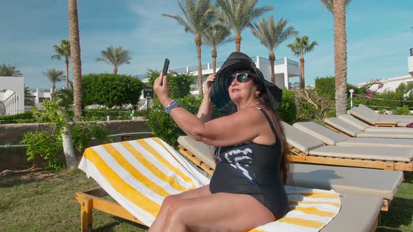 Caucasian Senior Female Elderly in a Black Hat Makes a Selfie on a Smartphone and Sunbathes