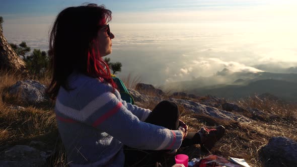 Young Redhead Woman in Sunglases Sitting on a Rock High in the Mountains Above the Clouds and