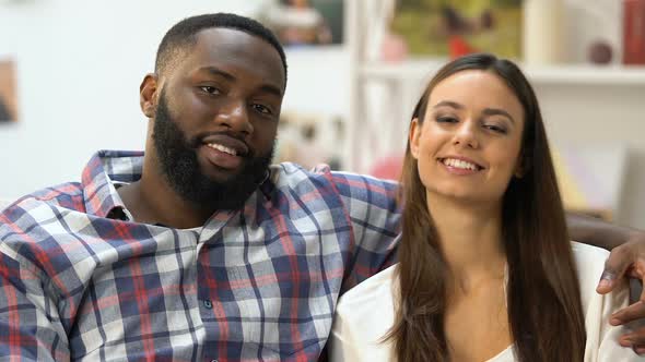 Beautiful Mixed-Race Couple Looking Into Camera and Hugging Sitting on Sofa