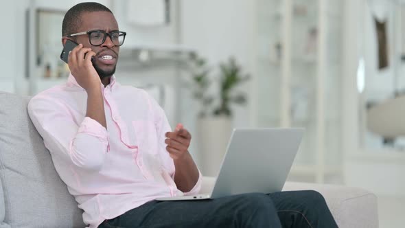African Man with Laptop Talking on Smartphone at Home
