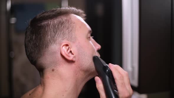 A man cutting his beard with a trimmer in front of a mirror