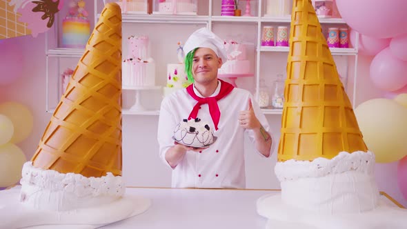 Young man dressed as chef holding cake, showing thumb up, standing in bright confectionery.