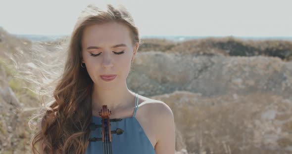 Elegant Woman Holds a Violin Opens Eyes and Looks Into Camera Among Cliffs