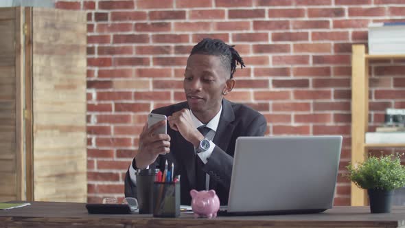 Black male businessman in office sits at a desk with a laptop and talks via video link using phone.