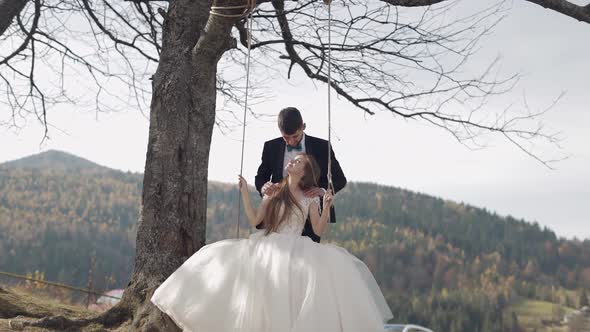 Newlyweds. Caucasian Groom with Bride Ride a Rope Swing on a Mountain Slope