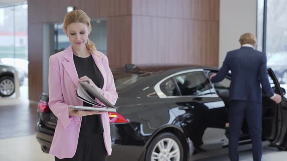 Portrait of Pleasant Cute Girl in Pink Jacket with a Big Book About Cars in Front of Couple Choosing