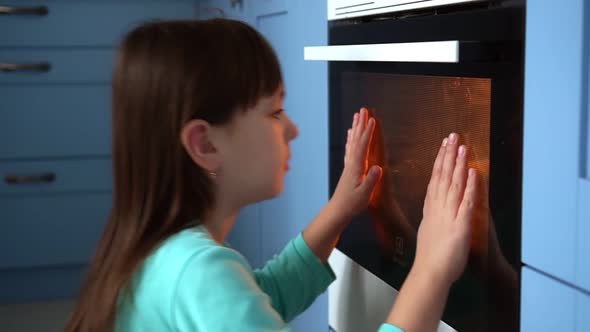 Cute Girl Sitting on Floor in Home Kitchen Looking at Oven and Waiting for Baking Food