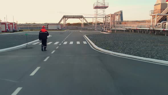 A Worker in an Orange Helmet Walks Along the Road Past Industrial Buildings and Pipelines