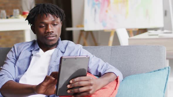 African american male artist smiling while using digital tablet sitting on the couch at art studio