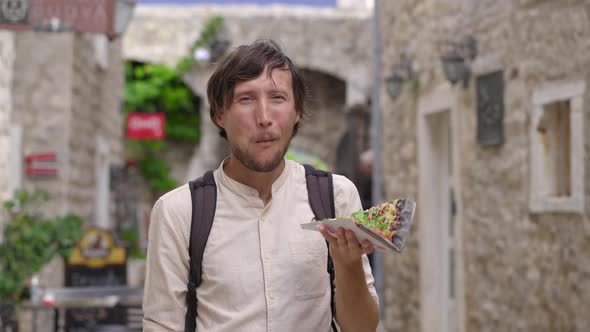 A Young Man Eats a Delicious Pizza on Black Bread Standing on a Street of an Old European Town