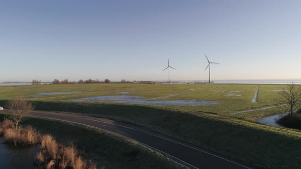 Aerial of White Car Driving on a Small Countryside Road with Two Windmills in the Background during