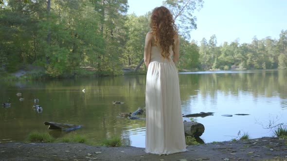 Back View of Beautiful Slim Woman with Red Hair in White Long Dress Standing on the Riverbank