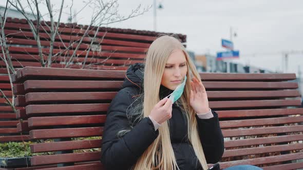 Young Woman Sitting on the Bench and Putting a Protective Medical Mask on Her Face