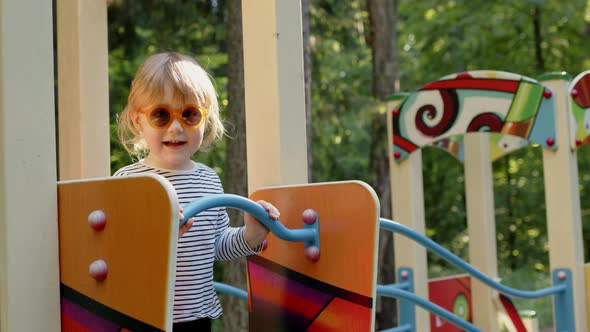 Little Cheerful Blonde Girl in Striped Tshirt and Red Sunglasses Stands on Playground and Nods Her