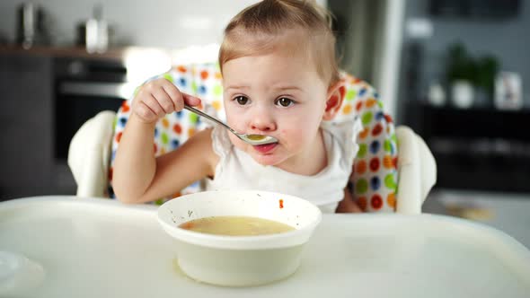 Cute Baby Girl Toddler Sitting in the High Chair and Eating Her Lunch Soup at Home Kitchen