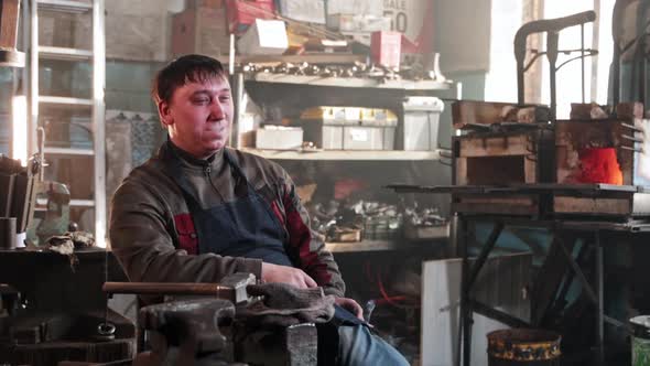 A Man Sitting in Blacksmith Workshop and Smoking a Cigarette and Looking in the Camera