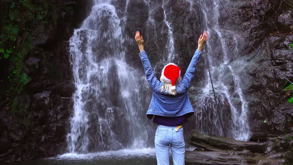 A girl in a Santa Claus hat stands on a waterfall with her arms outstretched.