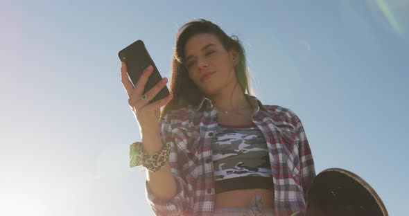 Smiling caucasian woman using smartphone and holding skateboard at a skatepark