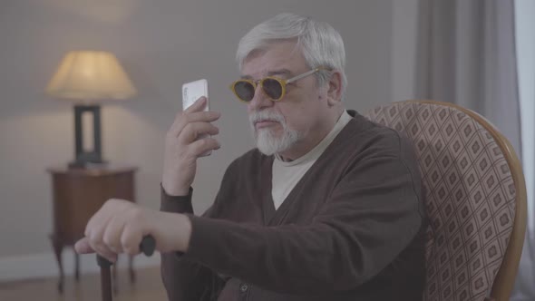 Portrait of Blind Old Caucasian Man Talking on the Phone Indoors. Grey-haired Male Retiree in