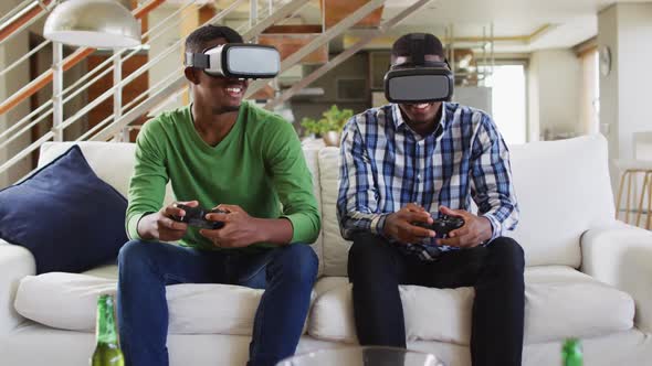 African american teenage twin brothers on couch using vr headsets and playing computer game smiling