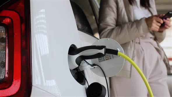 Charging of Electric Car Outdoors. Young Woman Waiting for Recharging of Eco Friendly Automobile and