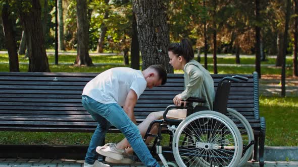 A Couple Walking in Park  a Guy Transfers Woman From a Wheelchair to Bench