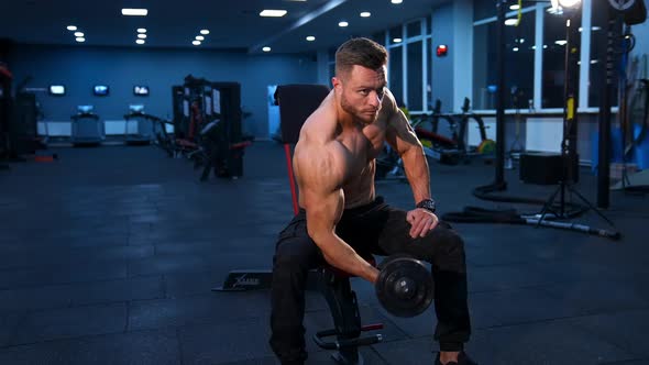 Power fitness man biceps. Handsome sportsman exercising with dumbbell while sitting in gym