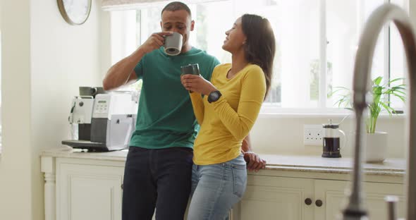 Happy biracial couple standing in kitchen, drinking coffee and laughing