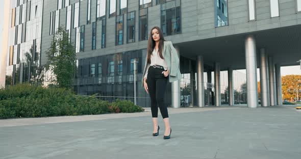 Beautiful Woman with Magnificent Figure Stands in Front of Modern Office Building Dressed in Elegant