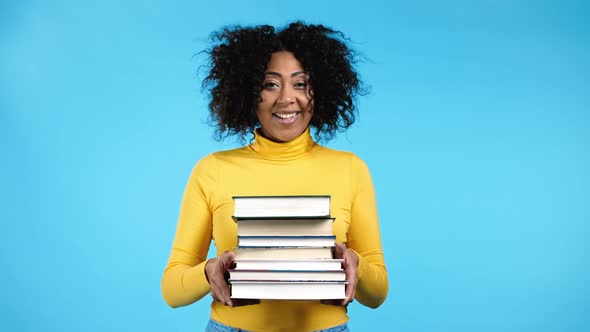 African American Student in Yellow Wear Holds Stack of University Books From Library on Blue