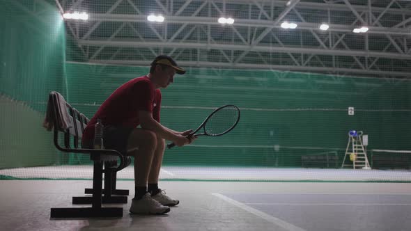 Male Tennis Player is Sitting on Bench in Tennis Court After Training or Match Resiting and Twisting