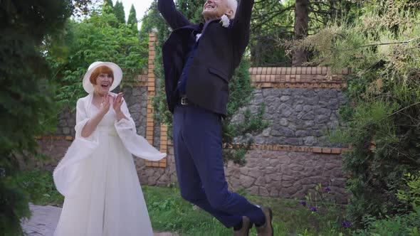 Excited Senior Groom Jumping in Slow Motion As Bride Clapping Standing Outdoors on Summer Spring Day