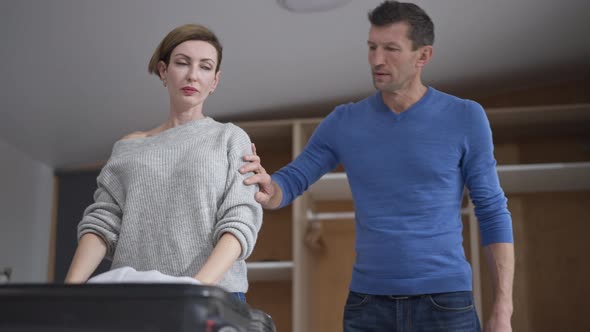 Sad Depressed Caucasian Husband and Wife Hugging As Woman Packing Suitcase at Home Indoors