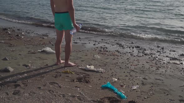 Unrecognizable Man Stands with His Back on Beach Looks Out to Sea with a Plastic Bottle in His Hands