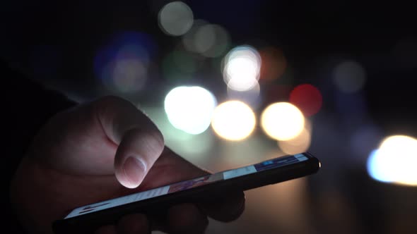 Hand of a Man With a Smartphone in the City at Night Against the Background of Blurry Lights of Cars